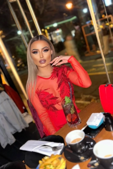 Elegant brunette escort in sheer red designer dress dining at London restaurant at night.