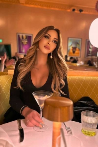 TS Bella Morgane is holding a glass sitting at a table 