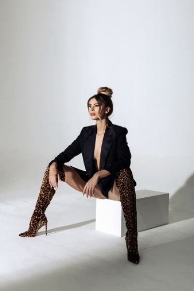 Blair is sitting on a box wearing a black blazer and knee high boots 