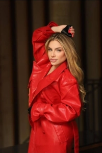 A blonde lady wearing a red leather coat and holding her hand above her head 