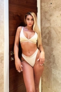 Blonde escort with a great figure standing against a wall 
