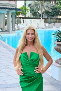 Blonde lady standing by a swimming pool wearing a green dress 