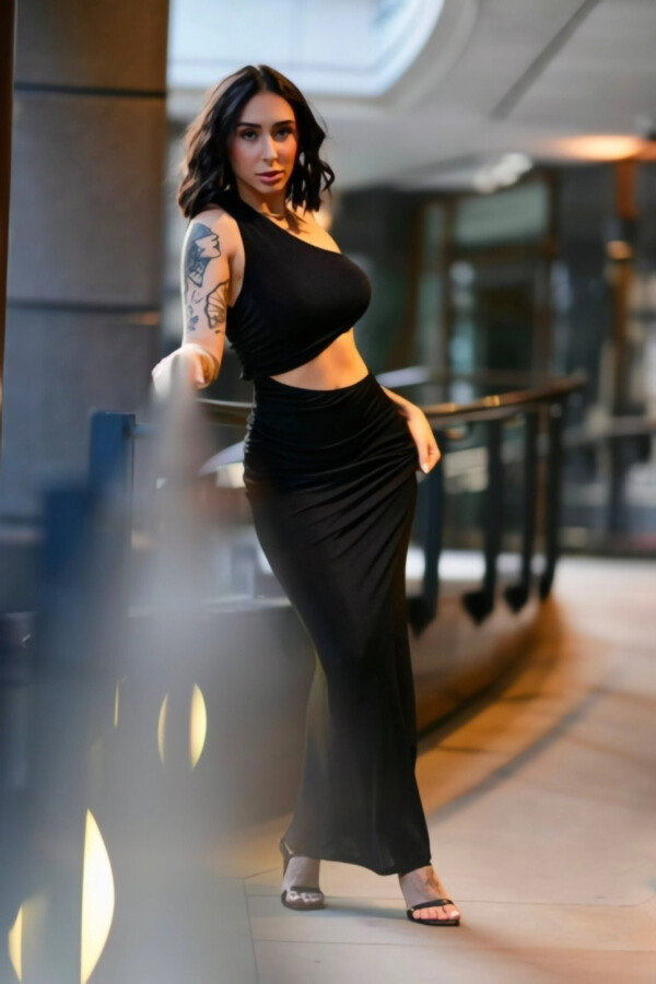Nay wearing a black one-shoulder dress on an outdoor walkway