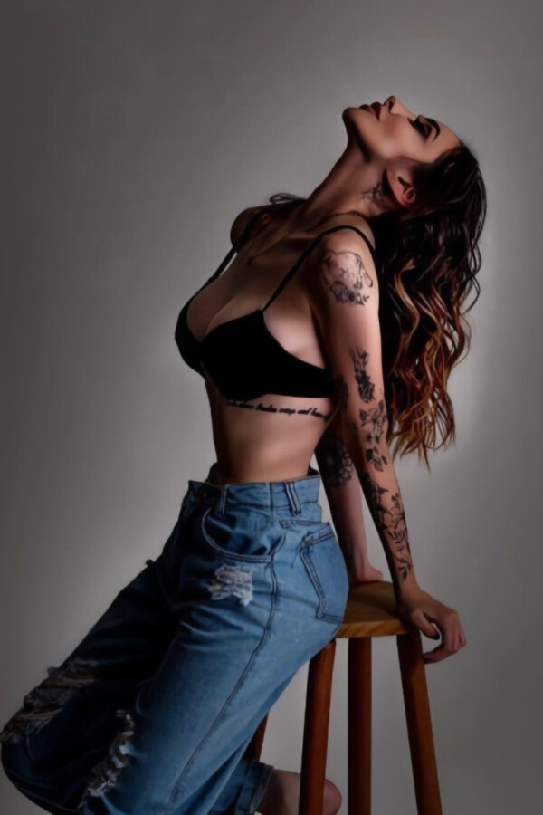 Rafaela leaning back on a wooden stool in a black bralette and jeans