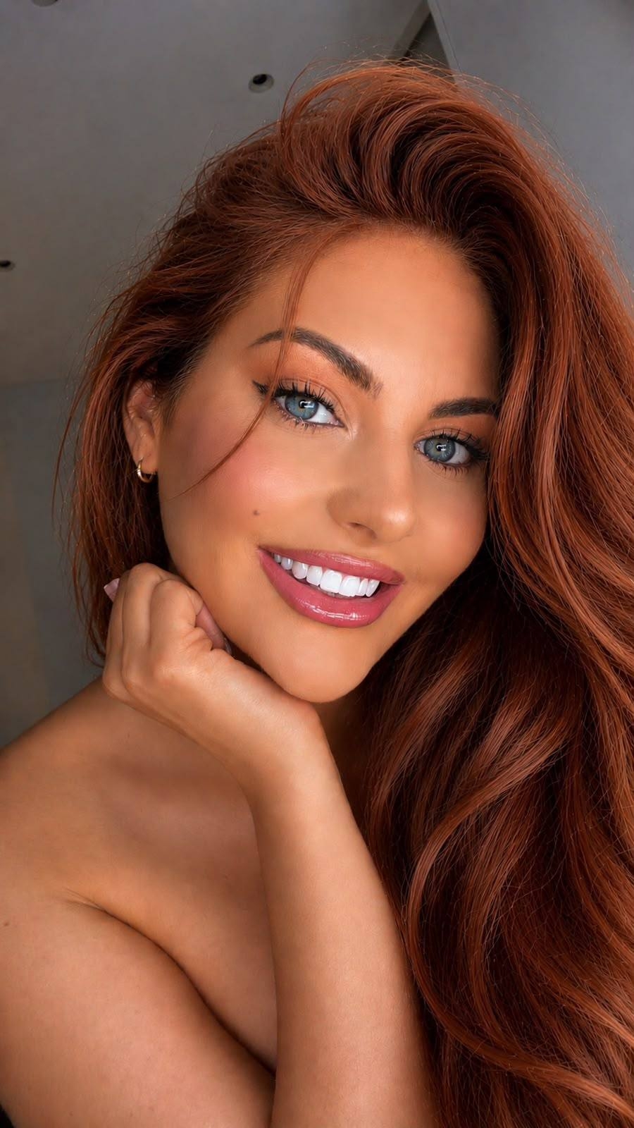 Aurora smiles at the camera in a close-up selfie with long auburn hair swept to one side.