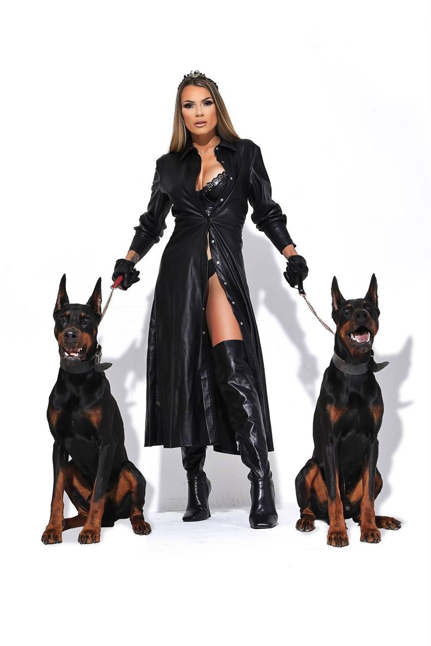 Mistress Loren stands between two Dobermans wearing a long black leather coat and knee-high boots.