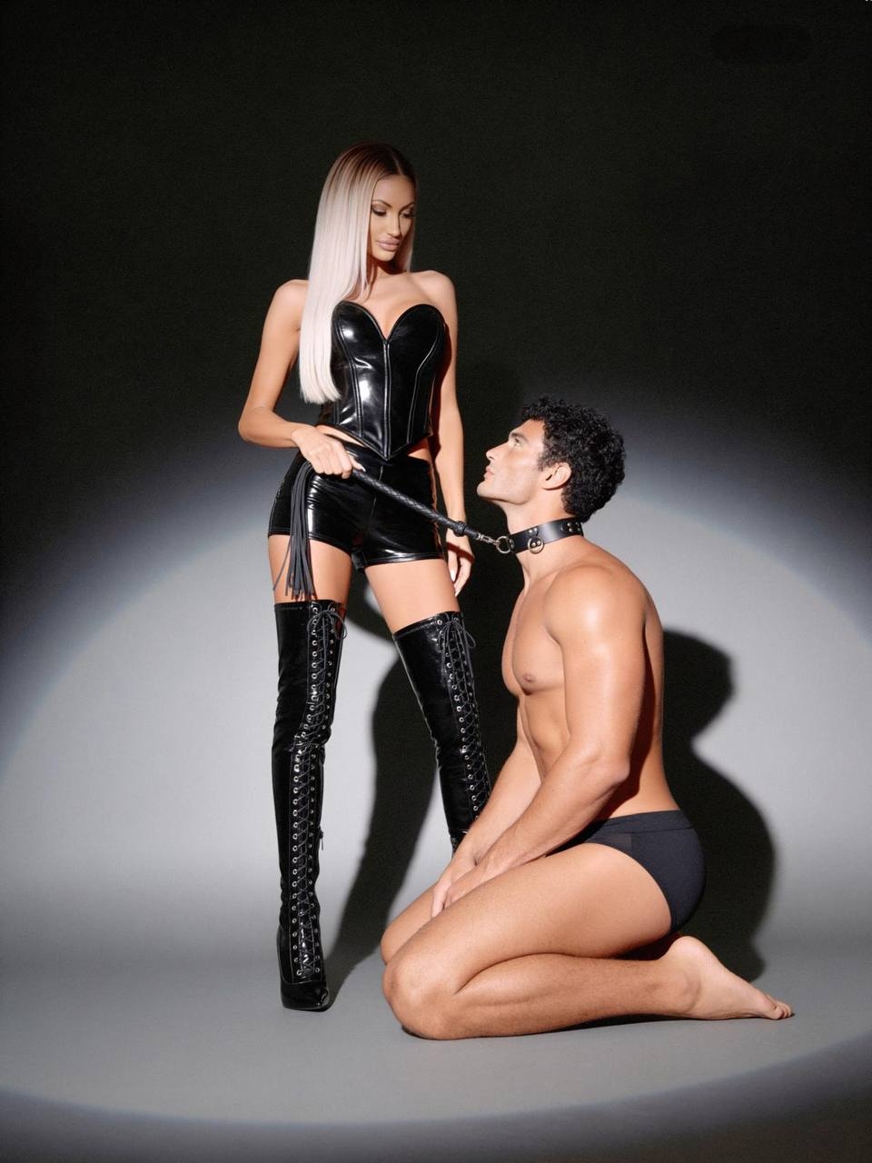 Mistress Bianca looks down at a kneeling man wearing a collar, holding his lead in one hand.